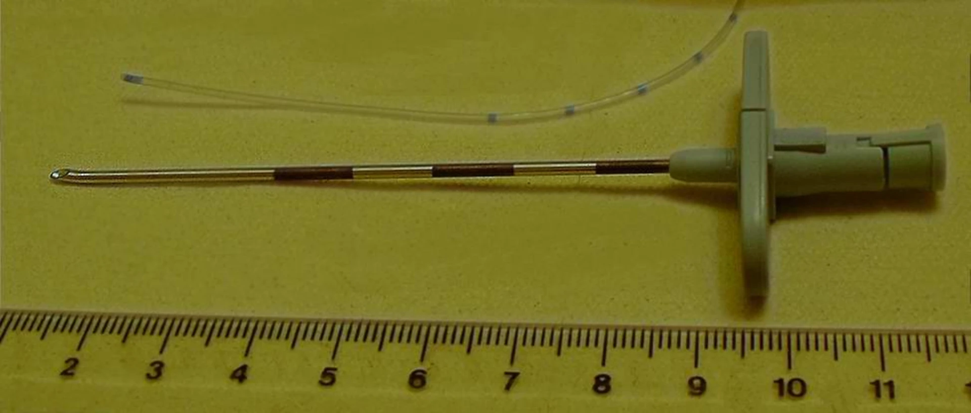 Tuohy epidural needle with stylet beside silicone catheter on calibrated ruler, illustrating the curved bevel tip and depth markings used for lumbar drain placement